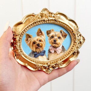 Hand holding an ornate gold oval frame containing a double pet portrait of two Yorkshire Terriers, one wearing a deep blue bow tie and the other a pale pink bow tie, set against a soft blue background.
