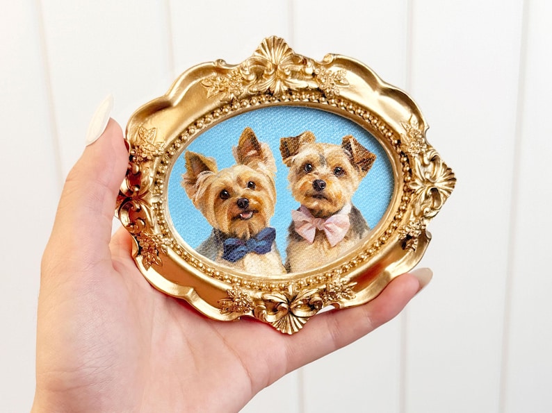 Hand holding an ornate gold oval frame containing a double pet portrait of two Yorkshire Terriers, one wearing a deep blue bow tie and the other a pale pink bow tie, set against a soft blue background.