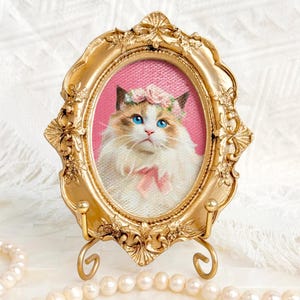 Portrait of a Ragdoll cat with a floral crown and pink bow, framed in an ornate vintage gold oval frame with a pink background and pearls around.