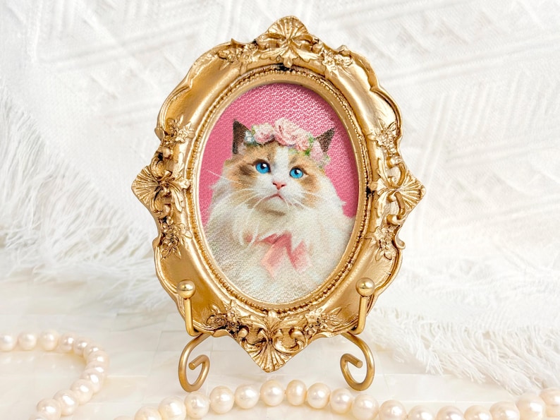 Portrait of a Ragdoll cat with a floral crown and pink bow, framed in an ornate vintage gold oval frame with a pink background and pearls around.