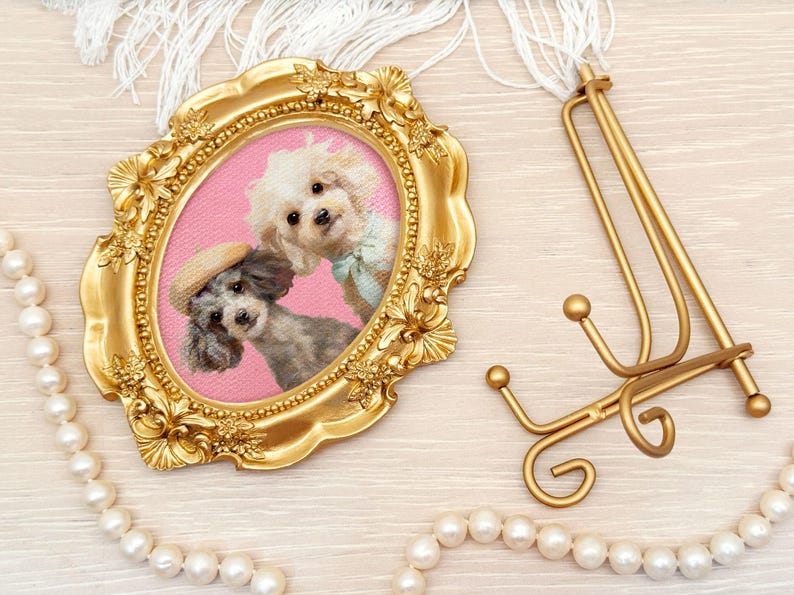 Acrylic painting of two poodles in vintage-style attire including a straw hat and green bow, displayed in an ornate gold frame with pearls and a wooden background.