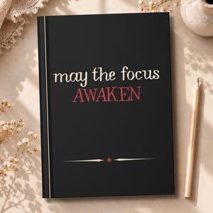 May include: A black hardcover journal featuring the white text "may the focus" above the red text "AWAKEN" and a small red diamond centered between two horizontal lines at the bottom of the front cover.