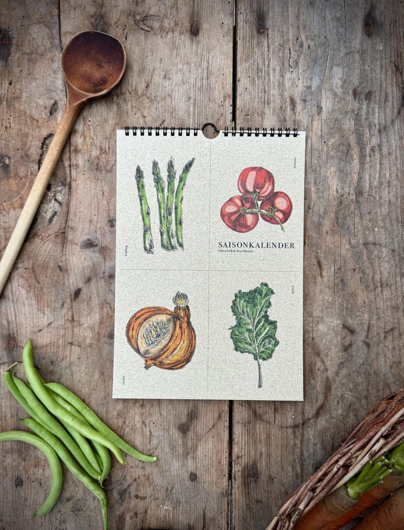 May include: A spiral-bound calendar with illustrations of vegetables, including asparagus, tomatoes, an onion, and kale. The calendar is open to a page with the German text "SAISONKALENDER". A wooden spoon and fresh green beans are also visible.