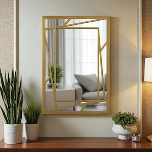 Puede incluir: Un espejo rectangular con un marco geométrico dorado. El espejo refleja una habitación con un sofá y cortinas. El espejo está colgado en una pared de color claro sobre una superficie de madera. Hay plantas en macetas en la superficie.