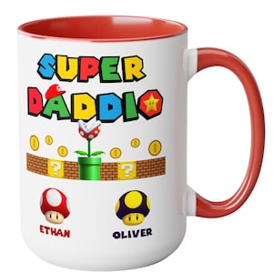 May include: White ceramic mug with a red interior and handle. The mug features the text "SUPER DADDIO" in colorful block letters, along with cartoon graphics of coins, mushrooms, and a Piranha Plant. The names "ETHAN" and "OLIVER" are printed below the mushrooms.