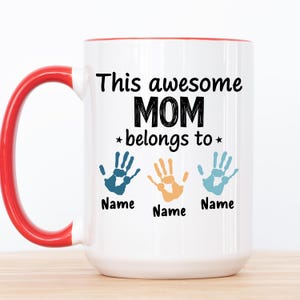 Peut inclure: Une tasse à café en céramique blanche avec un bord rouge. La tasse a un motif noir qui dit "This awesome MOM belongs to" avec trois empreintes de mains et le mot "Name" sous chaque empreinte de main.