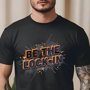Puede incluir: Camiseta negra con el texto "BE THE LOCK-IN" en naranja y azul, con un diseño geométrico. La camiseta está hecha de un material suave.