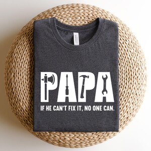 Könnte beinhalten: Ein dunkelgraues T-Shirt mit einer weißen Grafik, auf der "PAPA" mit Werkzeugen wie Hammer, Schraubendreher und Schraubenschlüssel steht. Der Text unter der Grafik lautet "Wenn er es nicht reparieren kann, kann es niemand."