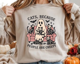 May include: Beige sweatshirt with a Halloween graphic. The design shows a ghost holding a cat, skeleton cats on a pink sofa, bats, and the text "CATS, BECAUSE PEOPLE ARE CREEPY."
