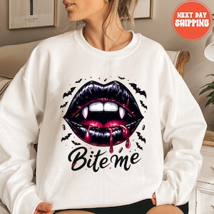 Könnte beinhalten: Weißes Sweatshirt mit einer Grafik von schwarzen Vampirlippen, Fangzähnen und tropfendem roten Blut. Das Design beinhaltet Fledermäuse und den Text "Bite me". Ein runder Aufkleber mit dem Text "NEXT DAY SHIPPING" ist sichtbar.