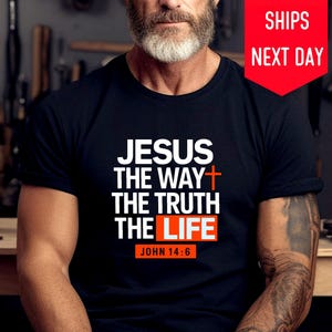 Puede incluir: Camiseta negra con el texto "JESUS THE WAY THE TRUTH THE LIFE" en blanco y naranja. El texto incluye un símbolo de cruz. La parte inferior de la camiseta dice "JOHN 14:6". Una pancarta roja en la esquina superior derecha dice "SHIPS NEXT DAY".