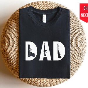 Pode incluir: Camiseta preta com a palavra "DAD" em letras brancas e uma silhueta de uma pistola, um rifle e uma pistola de cada lado das letras.