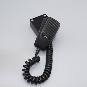 May include: Black handheld microphone with a coiled cord, attached to a black rectangular base. The microphone has a ridged front and the brand name "Johnson" is visible. The cord is tightly wound.