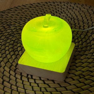 Puede incluir: Una lámpara brillante en forma de manzana verde, colocada sobre una base cuadrada de madera. La lámpara emite una luz verde neón brillante, y la base es de color marrón claro. Se encuentra sobre una estera redonda tejida.