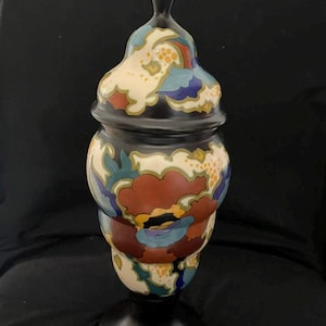 Può includere: Un alto vaso in ceramica con coperchio, caratterizzato da una base e un coperchio nero opaco con un pomello a punta. Il corpo e il coperchio sono decorati con un vivace motivo floreale multicolore nei toni del rosso scuro, blu, giallo e crema su fondo nero.