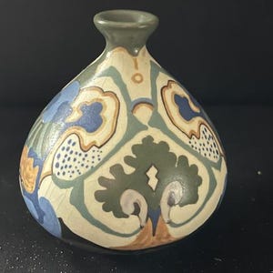 Op de afbeelding: Een decoratieve keramische vaas met een bolvormig lichaam en een smalle hals. De vaas heeft een crèmekleurige basis met een herhalend patroon van blauwe, groene en bruine bloemmotieven. De rand is een gedempt groene kleur.