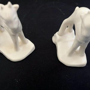 Puede incluir: Dos figuras de cerámica blanca de ciervos jóvenes, cada una sobre una pequeña base integrada. Las figuras están esculpidas con superficies lisas y mate, y presentan cuatro patas, una cola corta y orejas pequeñas.
