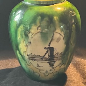 Può includere: Un piccolo vaso in ceramica verde con una scena dipinta in bianco e nero di un mulino a vento e una barca. Il vaso ha un bordo giallo.