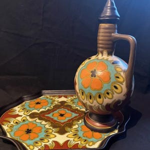 Op de afbeelding: Een keramische dienblad met een kleurrijk bloemenpatroon en een bijpassende keramische kan met een deksel. Het dienblad en de kan zijn versierd met oranje, blauwe, gele en bruine bloemen.