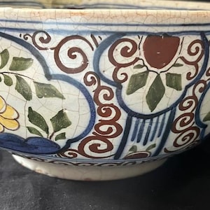 Può includere: Primo piano di una ciotola decorativa in ceramica con un motivo floreale bianco, blu e marrone. La ciotola presenta una glassa craquelé e un motivo ripetuto di fiori, foglie e volute. Il bordo è di colore crema chiaro.