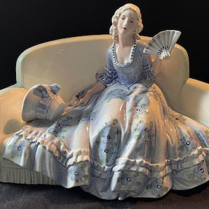 Op de afbeelding: Een porseleinen beeldje van een vrouw in een blauwe en witte jurk die op een witte bank zit. Ze houdt een waaier in haar rechterhand en heeft een hoed op haar schoot.