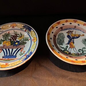 Può includere: Due piatti ovali in ceramica con disegni dipinti a mano. Un piatto presenta una composizione floreale in un vaso blu e bianco. L'altro piatto raffigura un uomo che suona il violino. Entrambi i piatti hanno bordi decorativi con accenti gialli, blu e marroni.