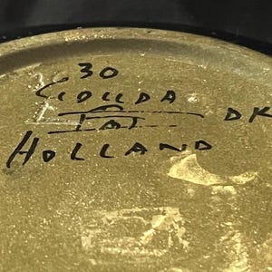 Puede incluir: Primer plano de la parte inferior de un objeto de cerámica con el texto escrito a mano "30", "HOLLAND" y "DK".