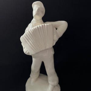 May include: A white ceramic figurine of a musician playing an accordion. The figure is wearing a cap and pants, standing on a round base. The accordion has a detailed, ribbed design. The figurine is set against a black background.