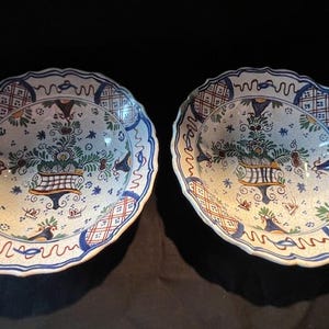 Può includere: Due piatti antichi con bordo smerlato e motivo floreale. I piatti presentano uno sfondo color crema con dettagli blu, verdi e arancioni. Il disegno centrale comprende una composizione floreale in un cesto.