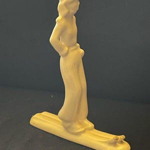May include: A cream-colored ceramic figurine depicting a skier standing on a single long ski. The figure wears a smooth, solid-colored outfit with a cap and has hands resting on the hips. The entire piece features a uniform matte finish.