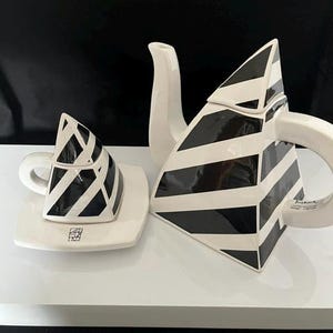 May include: A ceramic teapot and a matching cup with a saucer, both featuring a black and white diagonal striped pattern. The teapot has a triangular shape with a curved handle and spout. The cup is also triangular with a small handle and saucer.
