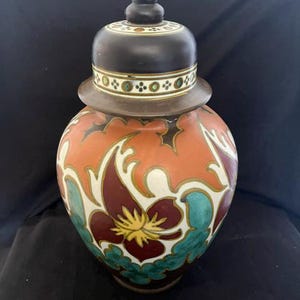 Op de afbeelding: Een keramieken gemberpot met afneembaar deksel, voorzien van een matzwarte afwerking op het deksel en de voet. Het lichaam toont een levendig patroon van gestileerde kastanjebruine, turquoise en gele bloemmotieven op een oranje en witte achtergrond.