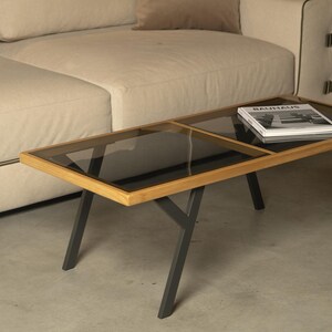 Puede incluir: Una mesa de centro moderna con diseño rectangular. Cuenta con un marco de madera, una tapa de vidrio oscuro y patas de metal gris oscuro. Un libro está colocado sobre la mesa. La mesa está frente a un sofá beige.