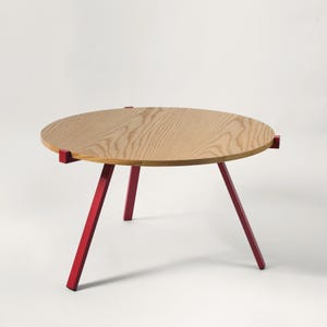Puede incluir: Una mesa de centro redonda de madera con un acabado de madera natural y patas de metal rojo. La mesa tiene un diseño simple y moderno.