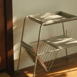Peut inclure: Une table d'appoint en métal gris clair avec deux étagères et un porte-revues. La table a une étagère supérieure rectangulaire et une étagère inférieure, avec un porte-revues en fil de fer. La table est sur un sol en bois.