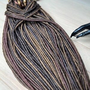May include: A bundle of brown, black, and light brown synthetic dreadlocks. The dreadlocks are arranged in a neat pile on a light wood surface.