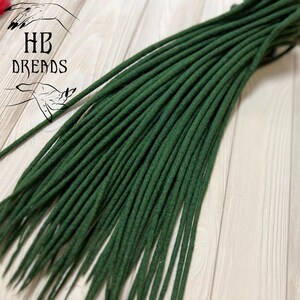May include: A bundle of dark green synthetic dreadlocks, arranged in a fan shape on a light wood surface. The dreadlocks are about 20 inches long.