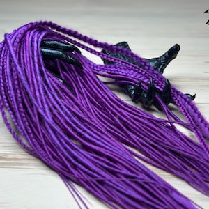 May include: Vibrant purple dreadlocks, partially braided, cascade from a black, claw-like sculpture. The image features the text "HB DREADS" with hand illustrations. The dreadlocks are a rich, saturated color, creating a striking visual contrast.