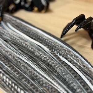 May include: A close-up of a bundle of synthetic dreadlocks in shades of gray and white. The dreadlocks are arranged in rows and are ready to be installed.