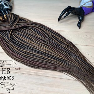 May include: A long set of brown, black, and burgundy synthetic dreadlocks. The dreadlocks are arranged in a loose braid and are lying on a wooden surface.