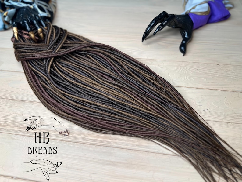 May include: A long set of brown, black, and burgundy synthetic dreadlocks. The dreadlocks are arranged in a loose braid and are lying on a wooden surface.
