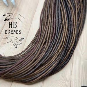 May include: A bundle of brown, dark brown, and burgundy synthetic dreadlocks. The dreadlocks are arranged in a loose pile on a wooden surface.
