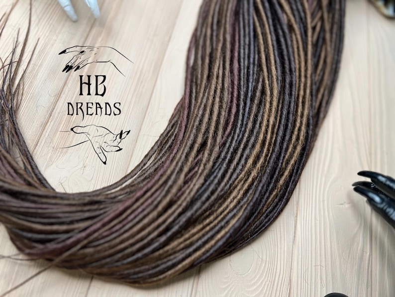 May include: A bundle of brown, dark brown, and burgundy synthetic dreadlocks. The dreadlocks are arranged in a loose pile on a wooden surface.