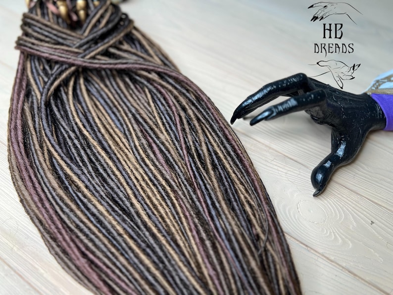 May include: A bundle of brown, black, and light brown synthetic dreadlocks. The dreadlocks are arranged in a neat pile on a light wood surface.
