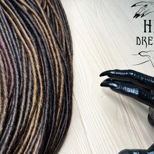 May include: A close-up of a bundle of synthetic dreadlocks in brown, dark brown, and black colors. The dreadlocks are arranged in a neat pile. The image also includes a black hand with long fingers and sharp nails. The hand is holding a sign that says "HB Dreads".