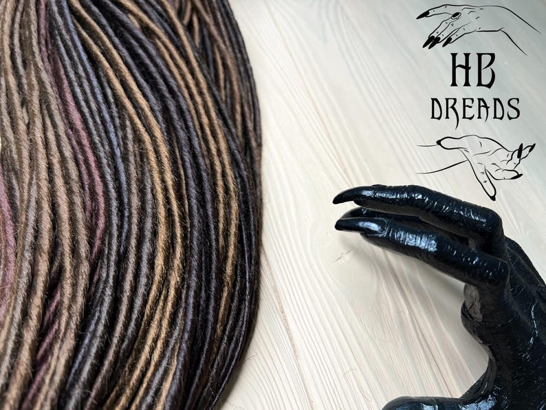 May include: A close-up of a bundle of synthetic dreadlocks in brown, dark brown, and black colors. The dreadlocks are arranged in a neat pile. The image also includes a black hand with long fingers and sharp nails. The hand is holding a sign that says "HB Dreads".