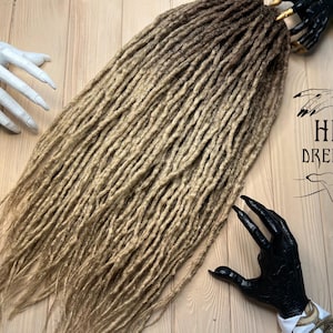 May include: A set of long, ombre synthetic dreadlocks in shades of blonde and brown. The dreadlocks are arranged in a loose, flowing style. The image includes the text "HB Dreads".