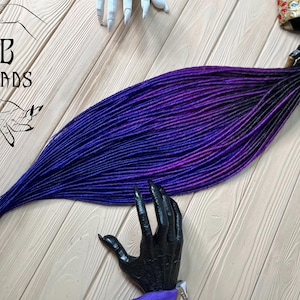 Puede incluir: Un conjunto de rastas sintéticas largas y de color degradado en tonos de morado, azul y negro. Las rastas están dispuestas en forma de abanico y se muestran sobre una superficie de madera.