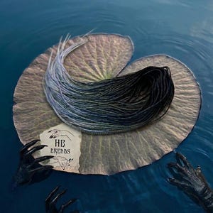May include: A set of synthetic dreadlocks featuring a color gradient that transitions from black at the roots to a light grey at the tips. The dreadlocks are coiled on a large lily pad, accompanied by a small paper card labeled HB DREADS.
