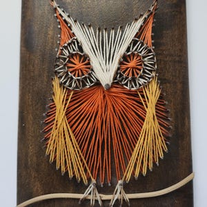 May include: A wooden plaque featuring a string art owl design. The owl is crafted with orange, yellow, and white thread, set against a dark brown background. The owl is perched on a thin, light brown branch.